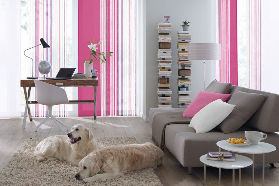 Zwei weiße große Hunde liegen auf einem flauschigen Teppich im Wohnzimmer. Im Hintergrund sind Streifengardine in Pink und weiß. Auf einem grauen Sofa liegt ein passendes pinkes Kissen zu den Gardinen.